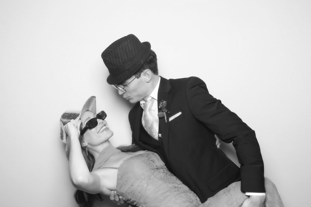 Black and white glam photo booth picture of a man in a fedora dipping a smiling woman in sunglasses