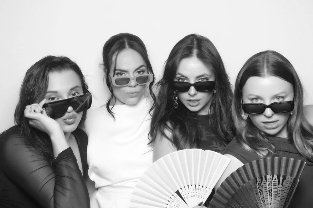 Black and white glam photo booth picture of four friends in sunglasses posing with hand fans