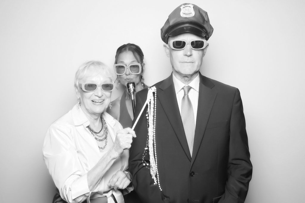 Black and white glam photo booth picture of three guests in sunglasses posing with a hat, microphone and pearl props